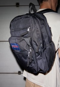 Mochila negra con múltiples compartimentos con cremallera, fabricada en un tejido duradero. Presenta un bolsillo lateral de malla y un parche del logo de Jansport en rojo y azul.