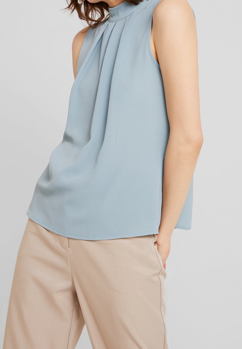 Blusa senza maniche azzurro chiaro con colletto alto, design frontale plissettato e texture liscia, abbinata a pantaloni beige.