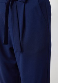 Mörkblå byxor gjorda av mjukt tyg, med ett knytbälte och sidofickor. Slät textur med en skräddarsydd passform.