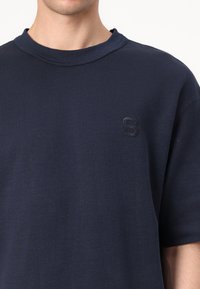 Tums zilas īsām piedurknēm T-krekls, izgatavots no kokvilnas, ar apaļu kaklu un diskrētu reljefā "S" logo uz krūtīm. Gluda tekstūra.