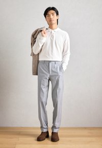 Ελαφρύ λευκό πόλο μπλουζάκι με μακριά μανίκια, γκρι παντελόνι με ρίγες και καφέ μποτάκια μέχρι τον αστράγαλο. Το μοντέλο κρατά μια μπεζ ζακέτα στον ώμο του.