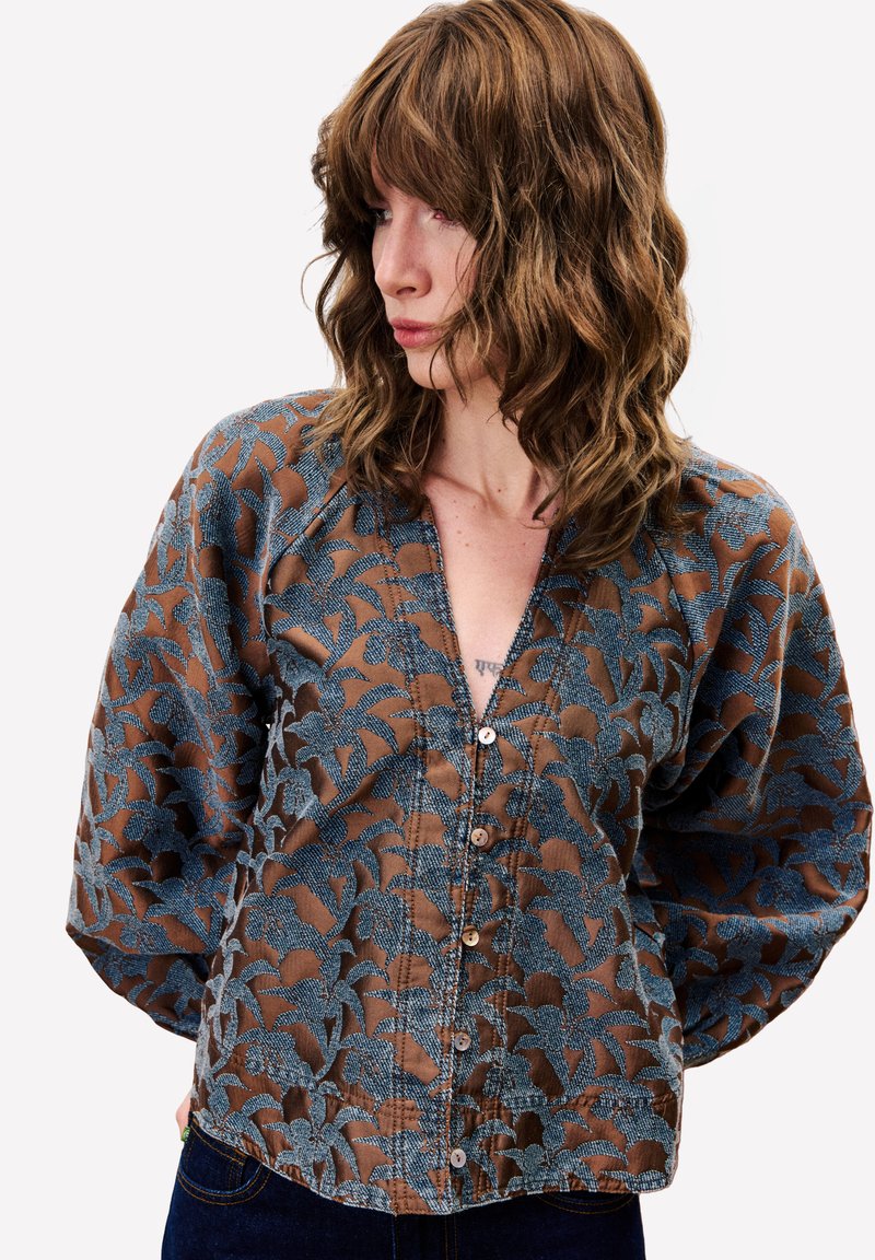 Frau mit welligem braunem Haar schaut nach links, trägt eine braune Bluse mit Knöpfen und blauem Blumenmuster sowie dunkle Jeans.