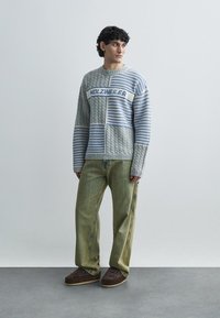 Pulover tricotat cu dungi gri și albastre, cu textură cable knit, având logo-ul "HOLZWEILER". Asortat cu pantaloni evazați din catifea bumbac verde deschis.