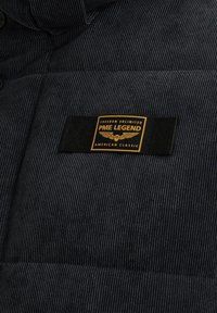 Tela de pana negra con un diseño acolchado. Presenta una etiqueta rectangular negra con texto amarillo que dice "PME LEGEND" y elementos decorativos.