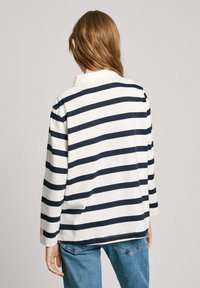 Weißes Langarmhemd mit dunkelblauen horizontalen Streifen, ausgestattet mit einem Kragen und einem lockeren Schnitt. Getragen mit blauen Jeans, was ein lässiges Design zeigt.