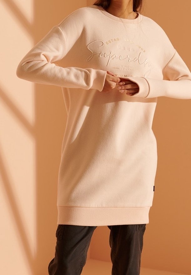 Femme portant une robe sweat à manches longues beige clair avec le texte "Superdry" en relief, ajustant les manches, debout devant un fond beige chaud.