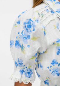 Vit blus med blå blommönster, puffärmar, plisserade detaljer och spetsdetaljer vid ärmsluten. Mjuk, lätt tygtextur.