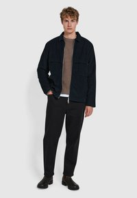 Dunkelblaues Cord-Jackett mit zwei Brusttaschen, über einem braunen ribbed Strickpullover und schwarzen Hosen getragen, kombiniert mit schwarzen Schuhen.