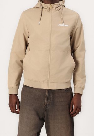 Mand iført beige lynlåshættejakke med "JACK & JONES"-logo på brystet og mørkebrune bukser, stående foran en ensfarvet baggrund.