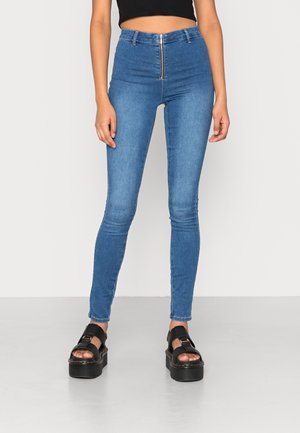 Jeans skinny azuis de brim de cintura alta com fecho na frente, apresentando uma textura suave e costuras mínimas. Combinados com sandálias de plataforma pretas.