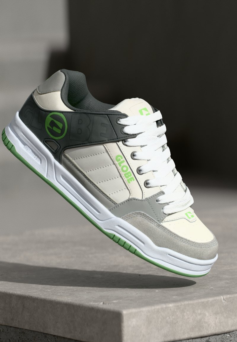Grå och creme sneakers med vita snören och gröna accenter. Har en gummisula, mocka och syntetiska material, samt en logotyp på sidan.