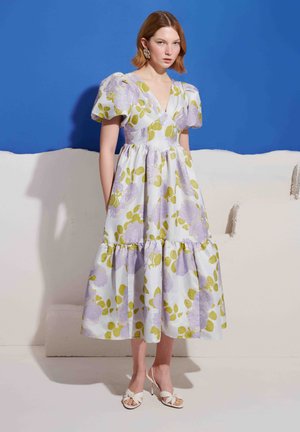 Femme debout devant un mur bleu et blanc, portant une robe midi blanche à manches bouffantes avec un imprimé floral vert et lavande, talons blancs.