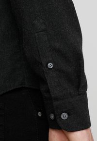 Zwart overhemd met knopen en gestructureerde stof. Het heeft lange mouwen, twee zwarte knopen aan de manchetten en een licht gebogen zoomlijn.