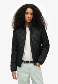 Zwarte gewatteerde jas met een hoge kraag, voorzien van een rits aan de voorkant en diagonale stiksels. Draagt over een wit T-shirt en lichte denim jeans.