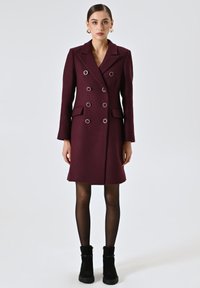 Cappotto bordeaux doppiopetto con hardware argento, rever a scollo a V, tasche laterali e silhouette aderente. Modella con stivali neri alla caviglia.