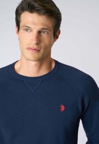 Maglione a maglia blu navy con scollo a girocollo, maniche raglan e un piccolo logo rosso sul petto. Texture morbida e design aderente.