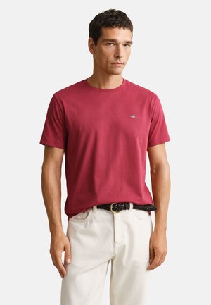 Hombre con una camiseta roja de cuello redondo y manga corta, pantalones blancos y un cinturón marrón trenzado, de pie frente a un fondo liso.