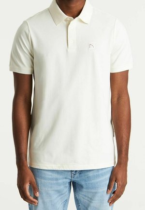 Man wearing a plain white short-sleeve polo shirt and light blue jeans, standing against a plain background.