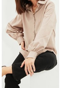 Chemise beige clair à boutons avec col et manches bouffantes ; dotée de poignets élastiques ; associée à un pantalon noir. Texture lisse, design minimal.