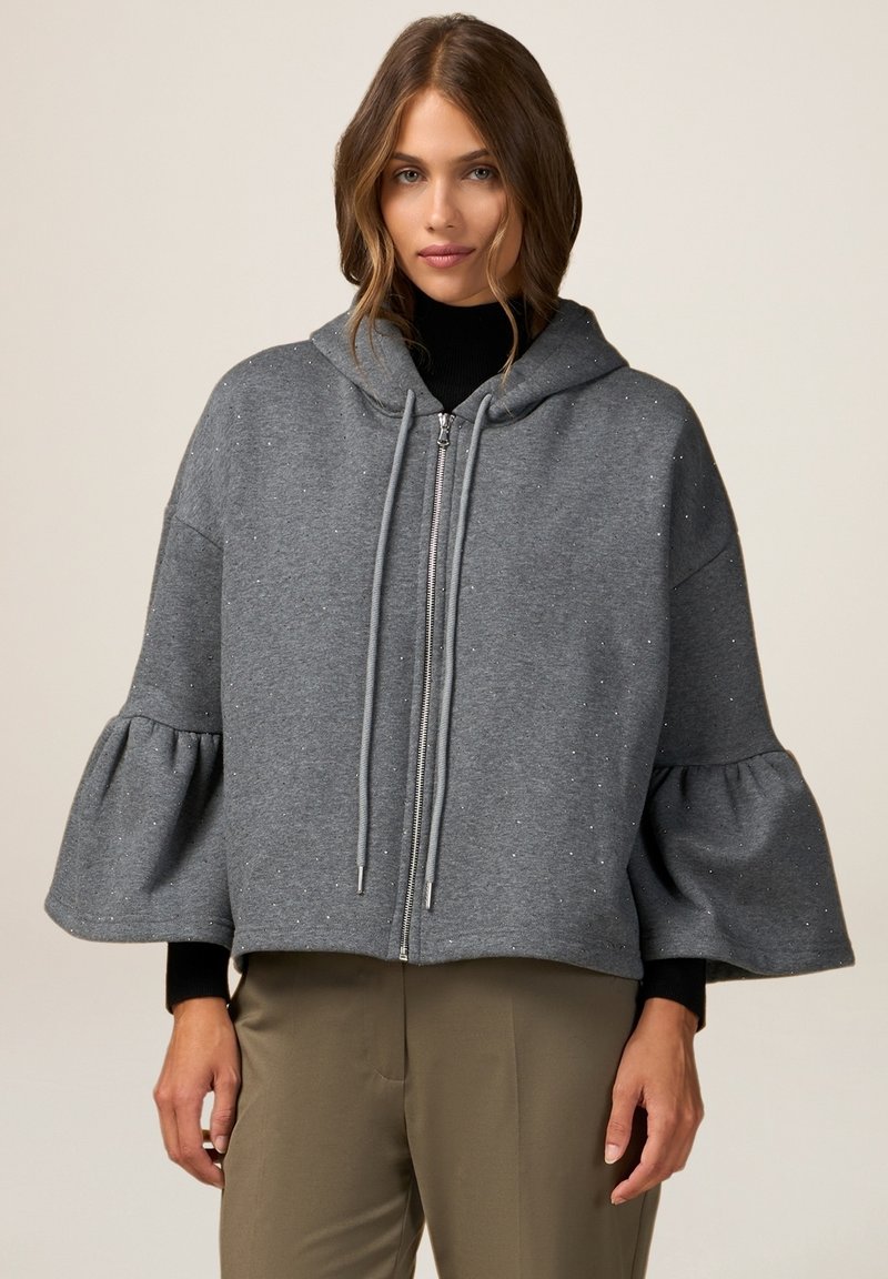 Hanorac gri cu fermoar, cu glugă, mâneci tip clopot și sclipici decorativ. Fabricat dintr-un material moale, cu o croială lejeră, se potrivește perfect cu un top și pantaloni negri.