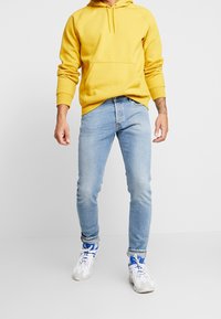 Gul hoodie, ljusblå jeans och vita sneakers med blå detaljer. Tyget verkar mjukt; jeansen är slim-fit med lätt slitningar.