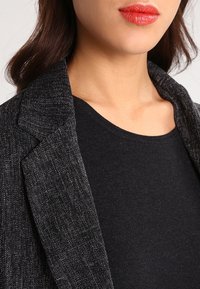 Femme portant un blazer texturé sombre et un haut noir, avec un rouge à lèvres rouge visible sur les lèvres, montrée du cou au haut de la poitrine.