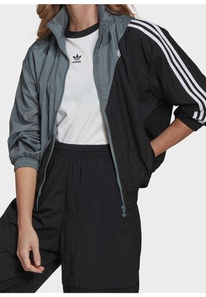 Osoba nosząca czarno-szarą kurtkę dresową Adidas z białymi paskami na rękawach oraz białą koszulkę z logo Adidas pod spodem.