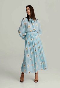 Robe bleu clair à imprimé floral, manches bouffantes longues, col à nouer, jupe plissée et sandales à talons noires. Tissu lisse et fluide.