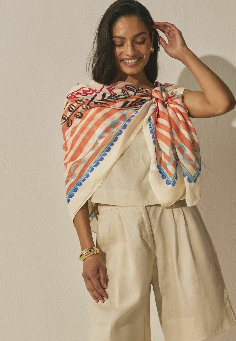 Sieviete smaida, valkājot krēmkrāsas apģērbu un krāsainu rakstainu šalli, kas sasieta plecā, ar zelta aproci un riņķa auskariem.