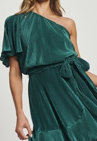Robe verte plissée, à une épaule, avec une taille à nouer. Son tissu doux et texturé renforce le mouvement et la tenue, avec des accents froncés.