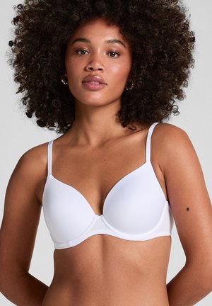 Woman with curly hair wearing a white padded bra with thin straps against a plain light gray background.