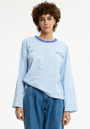 Mujer con cabello rizado que lleva una camisa de manga larga de rayas azul claro y vaqueros azules de talle alto, de pie frente a un fondo liso.