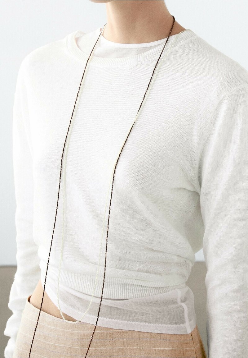 Witte, gebreide trui met een ronde hals en lange mouwen, gedragen over een doorschichtig topje. Geaccentueerd met een delicate, lange ketting.