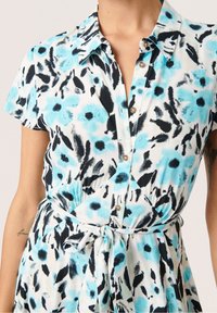 Košilové šaty z lehké látky s květinovým vzorem v modré, černé a bílé barvě. Mají límec, zapínání na knoflíky a vytažení v pase.