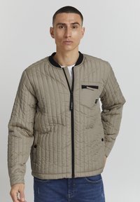 Giacca trapuntata beige chiaro con colletto nero e chiusura a zip. Presenta una tasca frontale con zip e dettagli con bottoni sui polsini.