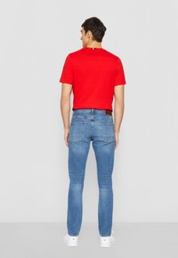 T-shirt rouge en coton à manches courtes, associé à un jean en denim bleu clair. Le jean est doté de poches arrière et d'une patch en cuir marron.
