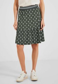 Grüner Stufenrock mit weißen Blumenmustern, elastischem Bund mit grauen Streifen, aus weichem Stoff. Kombiniert mit weißen Sneakern.