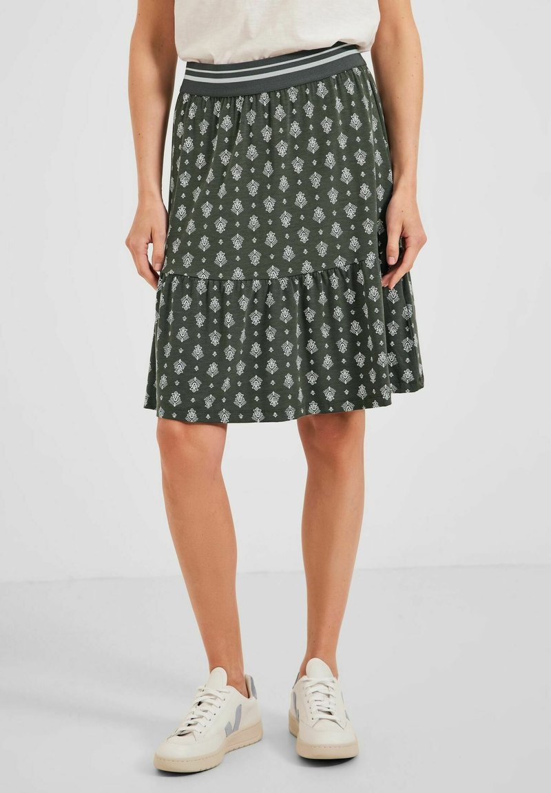 Grüner Stufenrock mit weißen Blumenmustern, elastischem Bund mit grauen Streifen, aus weichem Stoff. Kombiniert mit weißen Sneakern.