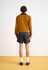 Brun långärmad topp med svart krage, i kombination med svarta mönstrade shorts. Modellen bär vita strumpor och svarta skor. Utsikt bakifrån.