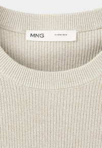 Pull beige en maille avec un col rond côtelé et une étiquette de taille MNG indiquant les tailles moyennes pour l’UE, les États-Unis et le Mexique.