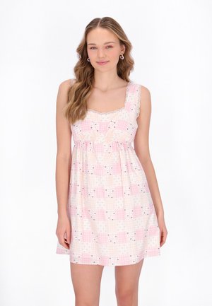 Ung kvinde med langt bølget hår iført en ærmeløs lyserød og hvid ternet kjole med blomsterprint, stående mod en hvid baggrund.