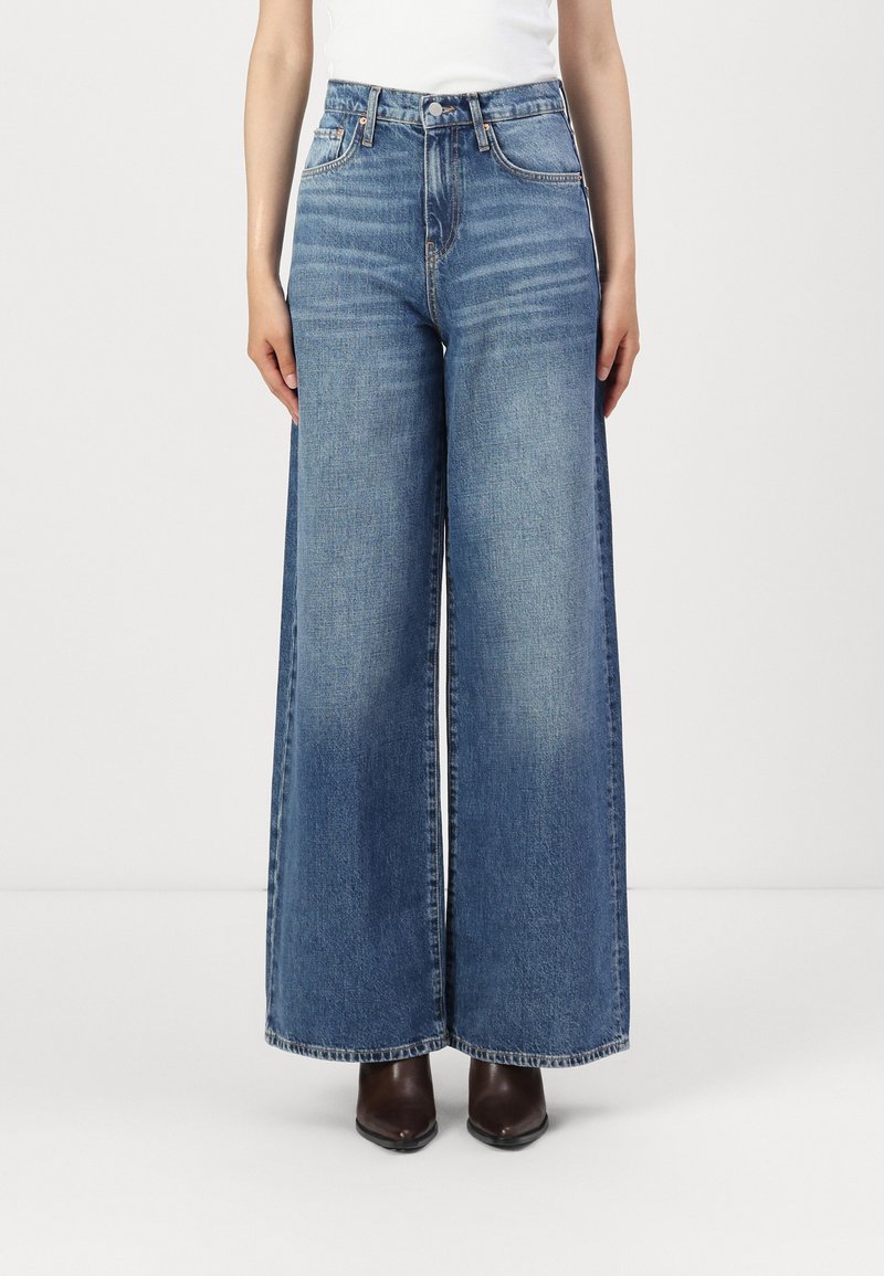 Jeans de pierna ancha en denim azul claro, con una cintura alta, un sutil desgastado y un diseño clásico de cinco bolsillos, combinados con botas oscuras hasta el tobillo.