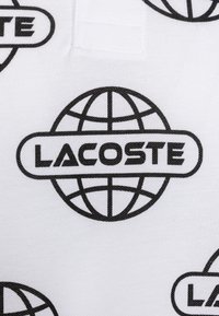 Vitt tyg med upprepade svarta globmönster och texten "LACOSTE", som visar en slät yta med en ren och modern design.