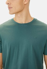 Turkis bomulds T-shirt med rund halsudskæring og korte ærmer. Har et lille broderet logo på venstre bryst. Glat tekstur.