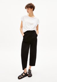 Zwarte hoge taille broeken met een losse pasvorm, gedragen met een losse witte overhemd met korte mouwen en zwarte sandaaltjes met bandjes. Neutrale achtergrond.