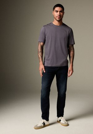 Muž s tetováním stojí, má na sobě šedé tričko s krátkým rukávem, tmavé džíny a bílé tenisky s černými pruhy na jednobarevném pozadí.