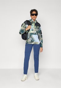 Kapa jakna s višeobraznim šumskim uzorkom, crnim akcentima, uparenim sa plavim hlačama i svijetlim cipelama, te crnim ruksakom.