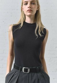 Haut sans manches noir avec un col montant, associé à un pantalon gris à rayures et une ceinture noire avec une boucle rectangulaire en argent.
