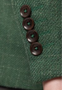 Zielona tkanina tweed z teksturowanym wzorem. Cztery ciemnobrązowe guziki z zielonymi detalami na rękawie.