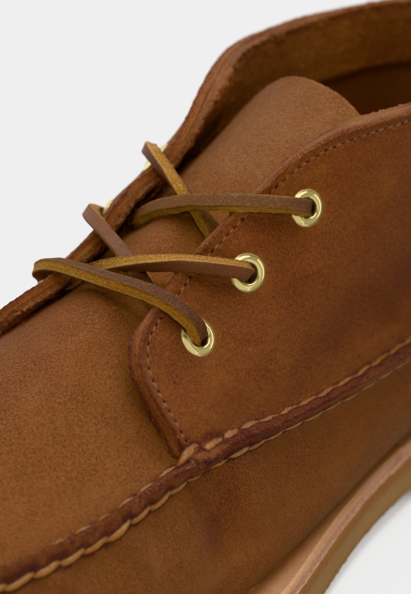 Scarpa in pelle marrone con una texture liscia, dotata di occhielli dorati e lacci abbinati, cuciture rinforzate lungo il bordo e un design semplice.